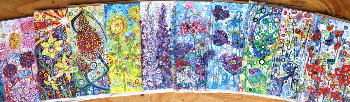 A set of colorful, hand-painted art cards or prints arranged in a fan shape on a wooden surface. Each card features vibrant, detailed floral and nature illustrations in a variety of colors including blue, yellow, red, green, and purple.