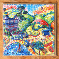 A square framed artwork featuring a highly colorful, whimsical landscape illustration with swirling circles, trees, hills, houses, and boats. The artwork is presented in a natural wood frame against a matching wood surface.